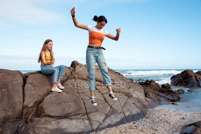 Вид спереди кавказца и смешанных расовых девушек, веселящихся вместе в солнечный день, одна девушка сидит на камне на пляже, другая прыгает с него. — стоковое фото