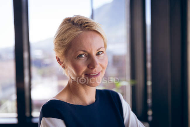 Retrato de una mujer de negocios caucásica, trabajando en una oficina moderna, de pie junto a una ventana, mirando a la cámara y sonriendo, en un día soleado - foto de stock
