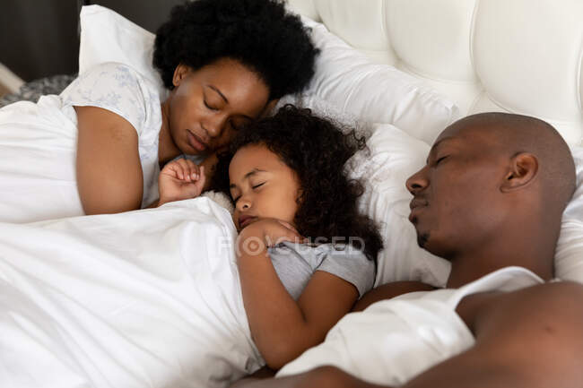 Высокий угол обзора афроамериканской пары и их юной дочери в спальне, лежащей вместе в постели, спящей — стоковое фото