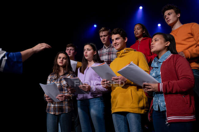 Вид спереди на многоэтническую группу юношей и девушек-хористов, держащих ноты и поющих стоя на сцене школьного театра во время репетиций спектакля, с рукой женщины-дирижера на переднем плане — стоковое фото