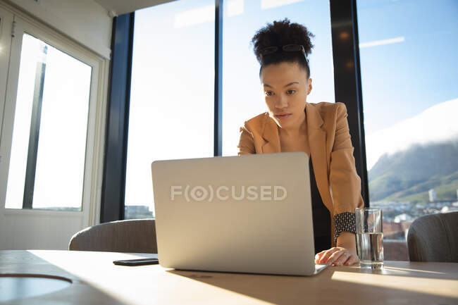Une femme d'affaires métisse travaillant dans un bureau moderne, debout près d'un bureau et utilisant un ordinateur portable, par une journée ensoleillée — Photo de stock