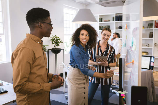Grupo multiétnico de colegas de negocios hombres y mujeres que trabajan en una oficina moderna, mirando una pizarra y hablando, con sus colegas trabajando en segundo plano - foto de stock