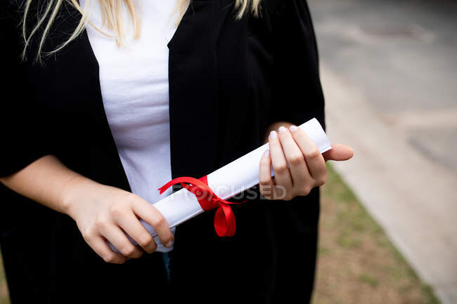 Visão frontal seção média de estudante do ensino médio feminino vestindo um vestido, segurando um diploma em seu dia de formatura — Fotografia de Stock