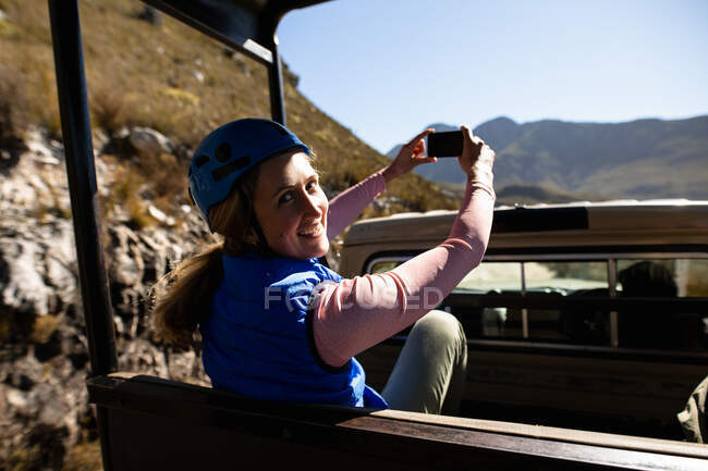 Portrait of Caucasian woman enjoying time in nature, in zip lining equipment sitting in a car, taking photos with smartphone on a sunny day in mountains — Stock Photo