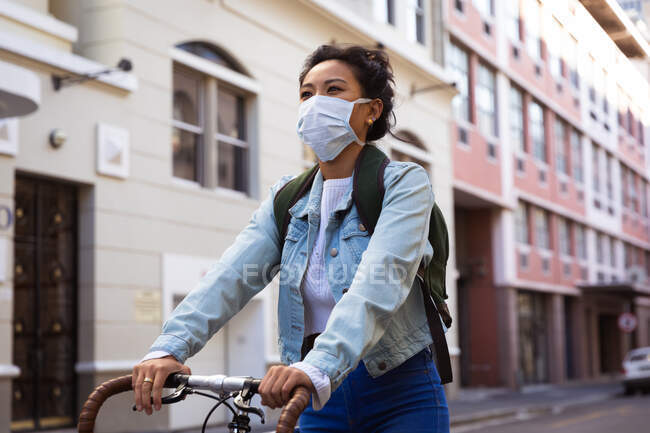 Низкий угол зрения смешанной расы женщина с темными волосами и вокруг на улицах города в течение дня, носить маску против загрязнения воздуха и коронавируса, езда на велосипеде со зданиями на заднем плане. — стоковое фото