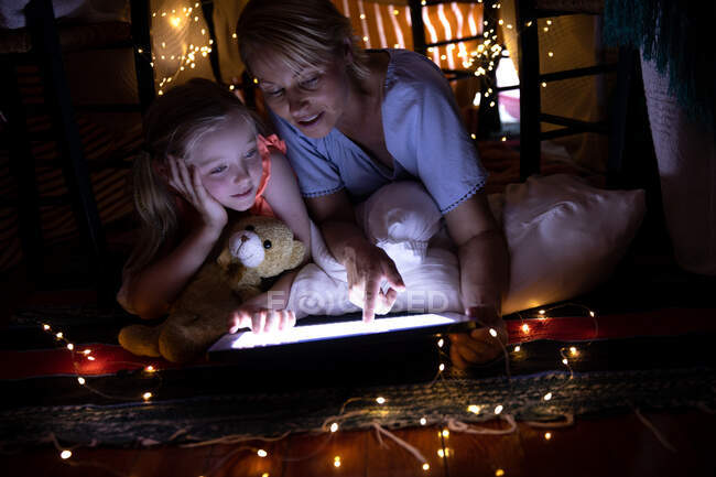 Vorderansicht einer kaukasischen Frau, die die Familienzeit mit ihrer Tochter zu Hause genießt, lächelnd in einem Zelt im Wohnzimmer liegend, mit digitalem Tablet, während ihre Tochter ihren Teddybär umarmt — Stockfoto