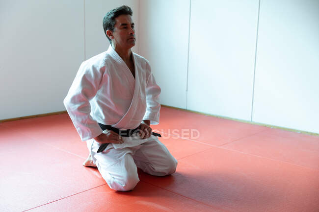 Vista frontal de um treinador de judô masculino de raça mista focado vestindo judogi branco, ajoelhado em esteiras no ginásio antes do treinamento de judô. — Fotografia de Stock