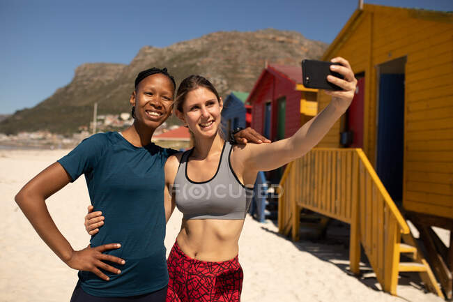 Vista frontal de mujeres de raza mixta disfrutando del tiempo en la playa soleada juntas, usando ropa deportiva, descansando después de trotar, mujer sosteniendo su teléfono inteligente, tomando selfie con su amigo. - foto de stock