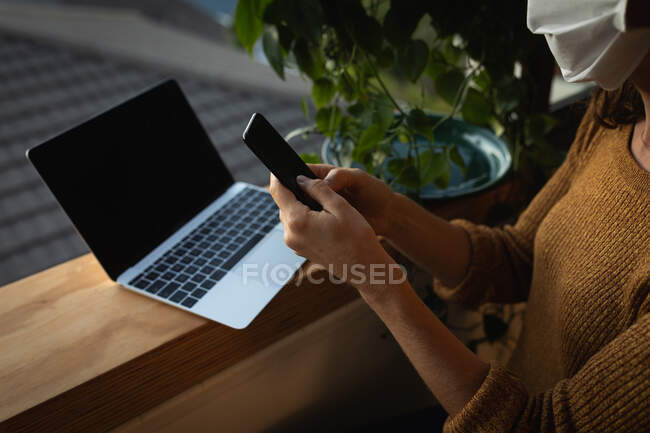 Mid section of a Caucasian woman spending time at home self isolating and social distancing in quarantine lockdown during coronavirus covid 19 epidemic, wearing a face mask, standing by a window, using her smartphone and a laptop computer. — Stock Photo
