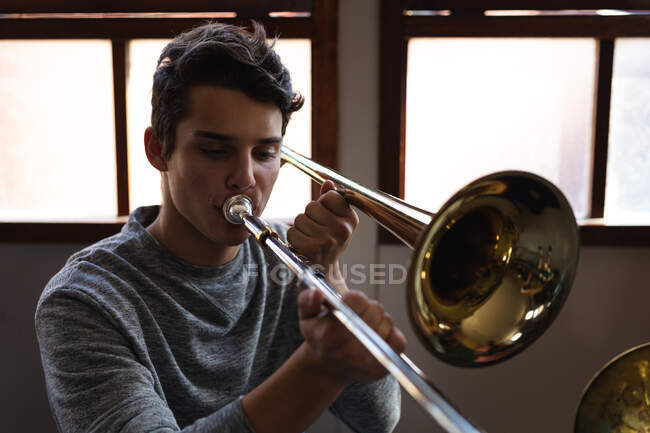 Vue de face gros plan d'un adolescent musicien caucasien avec un t-shirt gris assis devant une fenêtre jouant du trombone à l'école — Photo de stock