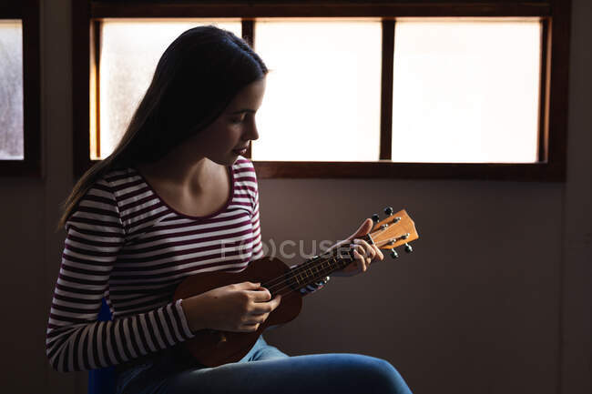 Seitenansicht einer kaukasischen Musikerin Teenager-Mädchen sitzt am Fenster und spielt eine Ukulele allein in einer High School — Stockfoto
