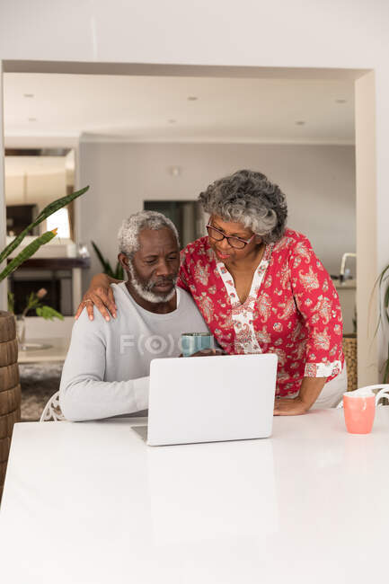 Una pareja afroamericana mayor pasa tiempo en casa juntos, distanciamiento social y aislamiento en cuarentena durante la epidemia de coronavirus covid 19, sentado en una mesa, usando una computadora portátil, abrazando - foto de stock