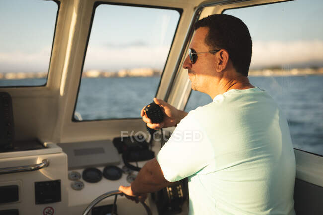 Ein kaukasischer Mann genießt seinen Urlaub in der Sonne an der Küste, auf einem Boot stehend, mit einem Walkie-Talkie — Stockfoto