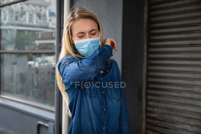Vista frontal de cerca de una mujer caucásica con máscara facial contra la contaminación del aire y covid19 coronavirus, caminando por la calle y cubriéndose la cara mientras tose. - foto de stock