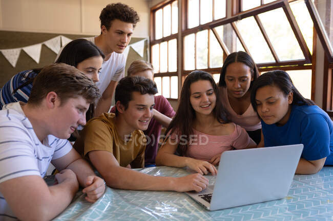 Vista frontal de un grupo multiétnico de alumnos adolescentes sentados en un aula mirando un ordenador portátil juntos y sonriendo en el descanso - foto de stock
