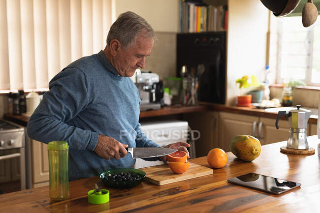 Vista lateral de un hombre caucásico mayor relajándose en casa, de pie en el mostrador en su cocina rebanando cuidadosamente la fruta por la mitad con un cuchillo afilado - foto de stock