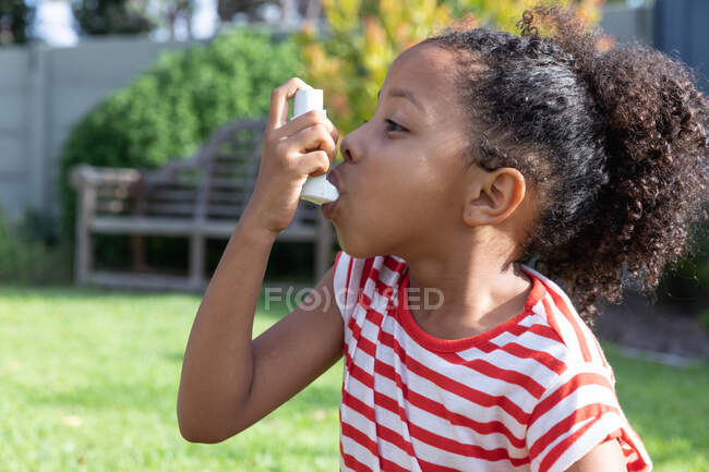 Афроамериканська дівчинка соціальна дистанція вдома під час карантину, одягнена в смугасту футболку, використовуючи її інгалятор, в саду в сонячний день.. — стокове фото