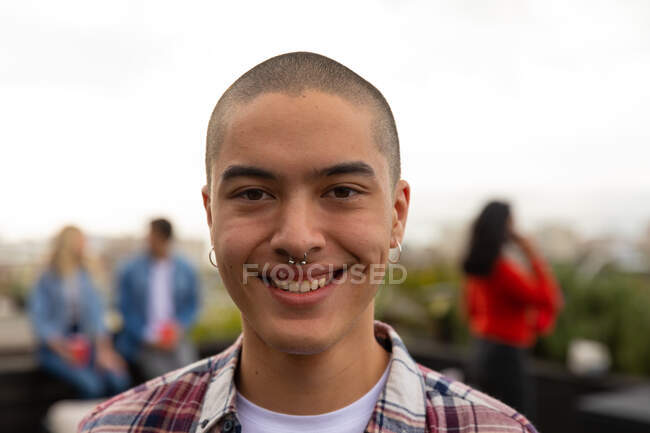 Portrait of a mixed race man hanging out on a roof terrace on a sunny day, looking at camera and smiling, with people talking in the background — Stock Photo