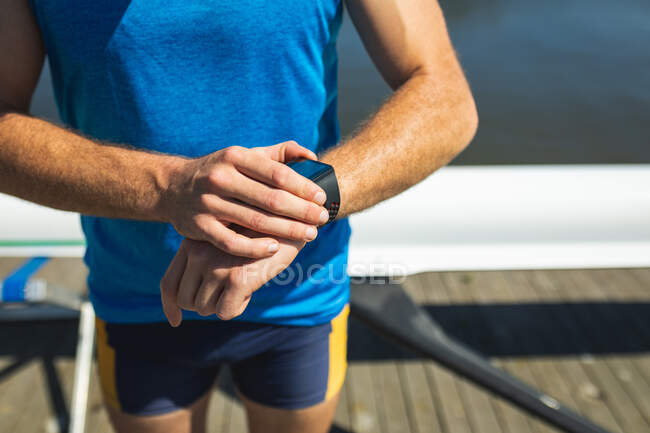 Frontansicht Mittelteil eines männlichen Ruderers, der auf seine Uhr blickt, auf einem Steg am Fluss stehend, im Hintergrund ein Ruderboot — Stockfoto