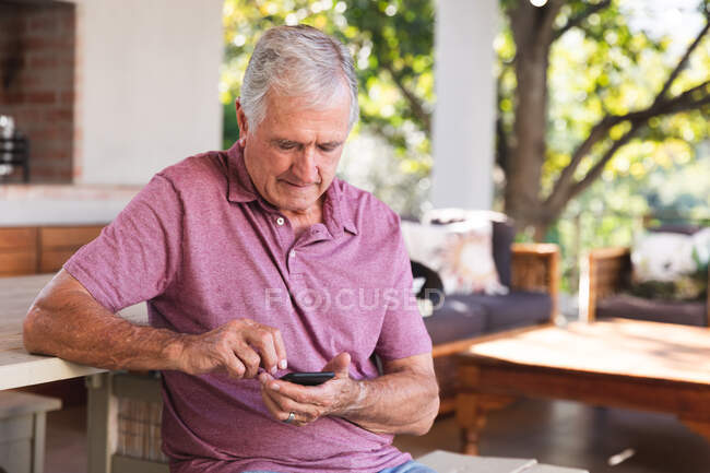 Красивый пожилой кавказский мужчина, наслаждающийся пенсией, сидящий за столом в саду на солнце, переписывающийся с мобильным телефоном, самоизолирующийся во время пандемии коронавируса — стоковое фото