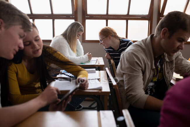 Side view of two teenage Caucasian girls talking in a school classroom, one turning around in her seat to talk to the other, sitting at a desk behind her, concentrating and writing — Stock Photo