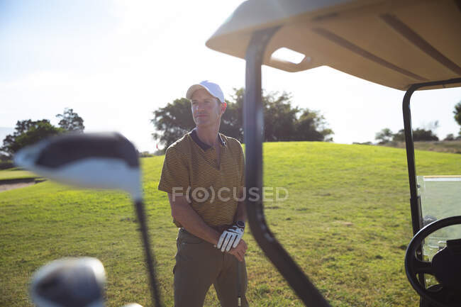 Vorderansicht eines kaukasischen Mannes auf einem Golfplatz, der an einem sonnigen Tag neben einem Golfwagen einen Golfschläger hält — Stockfoto