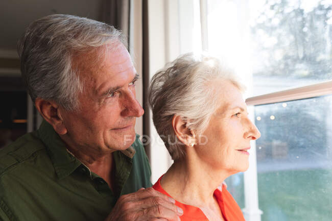 Happy retired senior Caucasian couple at home, embracing and smiling while looking out of the window together, couple at home together isolating during coronavirus covid19 pandemic — Stock Photo