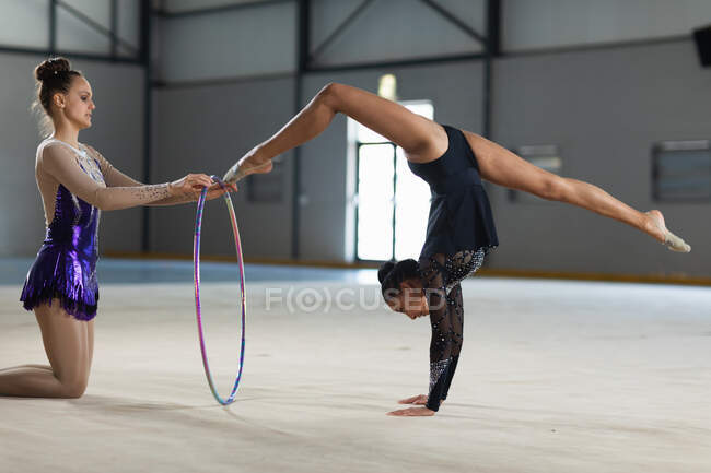 Side view of teenage Caucasian and mixed race female gymnasts performing at the gym, exercising with a hoop, girl in purple leotard holding the hoop, girl in black leotard doing handstand and split — Stock Photo