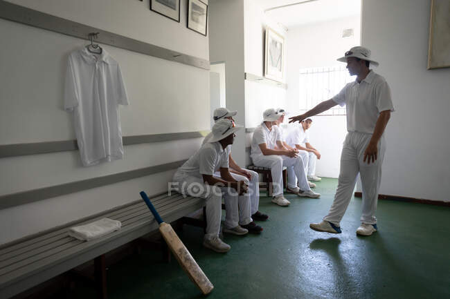 Seitenansicht eines Teams multiethnischer Cricketspieler, die Weiße tragen, auf einer Bank in einer Umkleidekabine sitzen und sich auf das Spiel vorbereiten, während einer der Spieler neben der Bank steht. — Stockfoto
