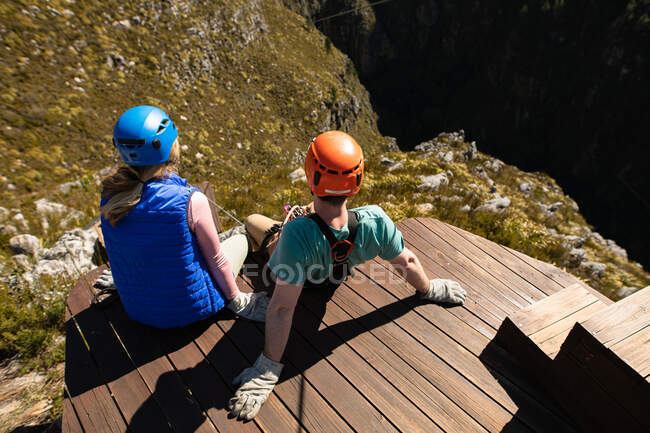 Vue arrière à angle élevé du couple caucasien profitant du temps dans la nature ensemble, portant un équipement de tyrolienne, assis sur le pont par une journée ensoleillée dans les montagnes — Photo de stock