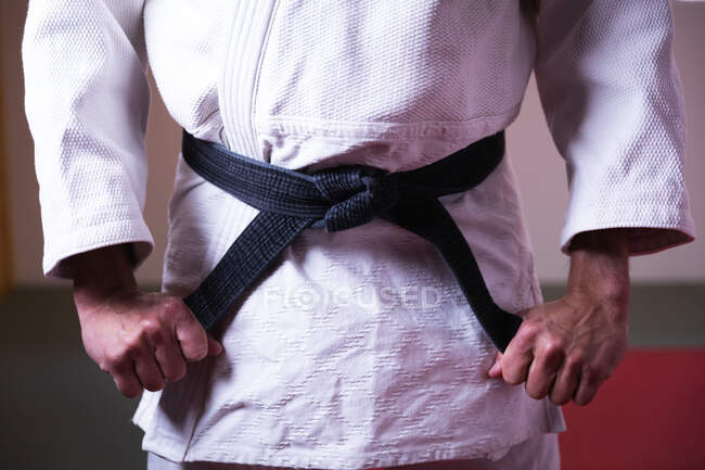 Front view mid section of judoka standing on gym mats, tying up the black belt of white judogi. — Stock Photo