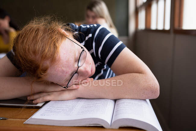 Seitenansicht eines jugendlichen kaukasischen Mädchens in einem Klassenzimmer der Schule, das mit dem Kopf auf den Armen an ihrem Schreibtisch sitzt und schläft, während männliche und weibliche Teenager an Schreibtischen im Hintergrund arbeiten. — Stockfoto