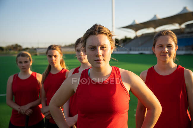 Portrait of a Caucasian female field hockey players, training before a game, standing on a hockey pitch, looking at camera, with her teammates standing in a row behind her on a sunny day — Stock Photo