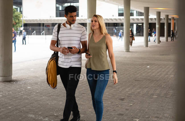 Vista frontal de una pareja caucásica por las calles de la ciudad durante el día, caminando, hablando y usando sus teléfonos inteligentes. - foto de stock