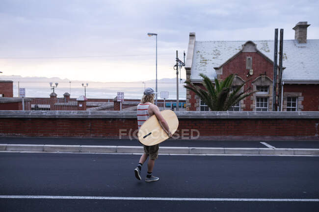 Vue arrière d'un surfeur caucasien aux longs cheveux blonds, vêtu d'une casquette pleine et de vêtements décontractés, tenant une planche de surf en bois et traversant la rue. — Photo de stock
