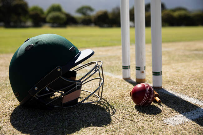Крупним планом вид на червоний крикетний м'яч і зелений шолом крикету, що лежить на крикетному полі поруч з хвірткою в сонячний день — стокове фото