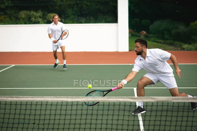 Ein kaukasischer und ein gemischter Rasse Männer in weißen Tennisanzügen verbringen Zeit auf einem Tennisplatz zusammen, spielen Tennis an einem sonnigen Tag, halten Tennis, einer von ihnen schlägt einen Ball — Stockfoto