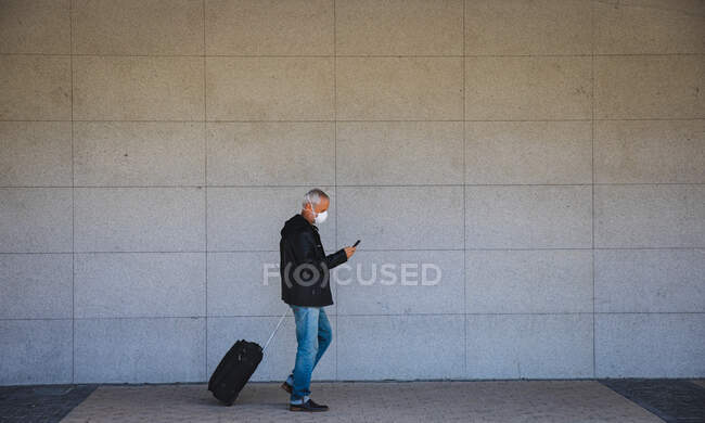 Старший кавказский мужчина днем бродит по улицам города в маске против коронавируса, ковид 19, использует смартфон и вытаскивает чемодан. — стоковое фото