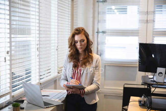 Negocio femenino caucásico creativo trabajando en una oficina moderna informal, de pie en un escritorio con una computadora portátil, con una chaqueta blanca, tomando notas - foto de stock