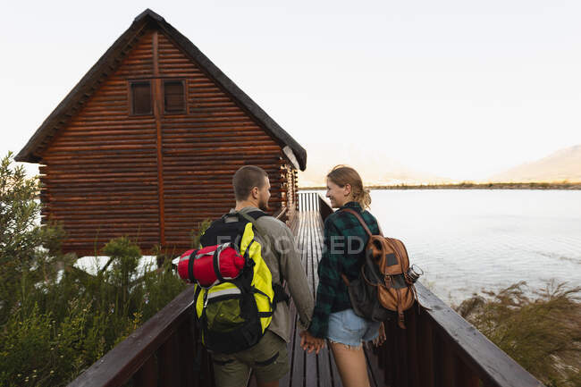 Vue arrière d'un couple caucasien s'amusant lors d'un voyage à la montagne, marchant sur un pont vers une cabane, se tenant la main — Photo de stock