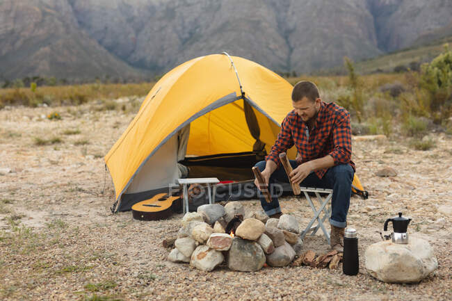 Vista lateral de un hombre caucásico pasándola bien en un viaje a las montañas, sentado junto a una fogata, sosteniendo un tronco y prendiéndolo fuego - foto de stock