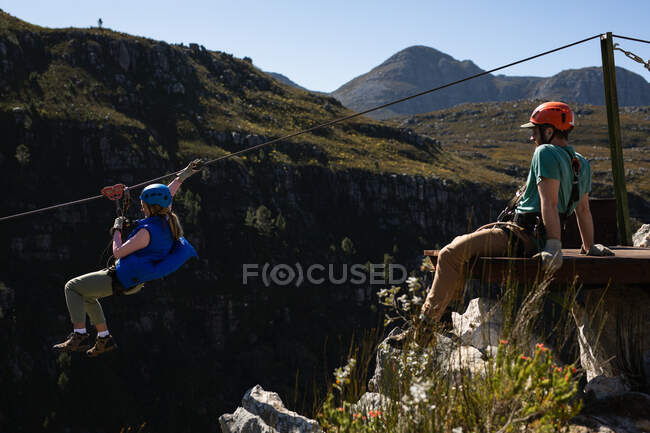 Side view of Caucasian woman enjoying time in nature together, zip lining while a young Caucasian man is watching her ride on a sunny day in mountains — Stock Photo
