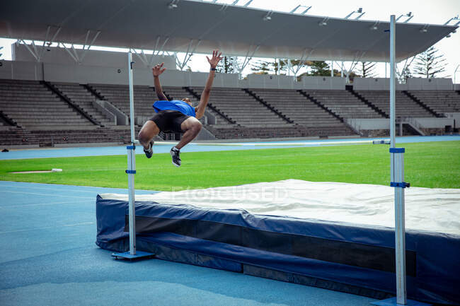 Vista frontal de un atleta masculino de raza mixta practicando en un estadio deportivo, haciendo un salto de altura. Entrenamiento deportivo de pista y campo en el estadio. - foto de stock
