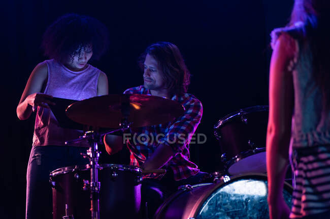 Front view of a Caucasian man sitting at a drum kit and a mixed race woman standing beside him, looking at a tablet computer together while setting up their band equipment on stage — Stock Photo