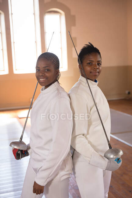 Portrait of two African American sportswomen wearing protective fencing outfits during a fencing training session, looking at camera and smiling, holding epees. Fencers training at a gym. — Stock Photo