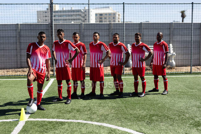 Équipe multiethnique de joueurs de football masculin portant une formation de bande d'équipe sur un terrain de sport au soleil, posant à la caméra avec un ballon. — Photo de stock