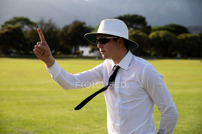 Vista lateral de un árbitro de cricket masculino caucásico con camisa blanca, corbata negra, un sombrero de ala ancha y gafas de sol, de pie en un campo de cricket en un día soleado, levantando la mano con un dedo hacia arriba. - foto de stock