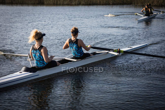 Rear view of two Caucasian female rowers from a rowing team training on the river, rowing in a racing shell on the water, with their teammates rowing in another boat in the background — Stock Photo
