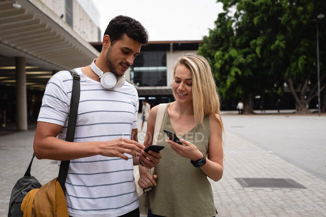 Vista frontal de una pareja caucásica por las calles de la ciudad durante el día, de pie en la calle y usando sus teléfonos inteligentes. - foto de stock