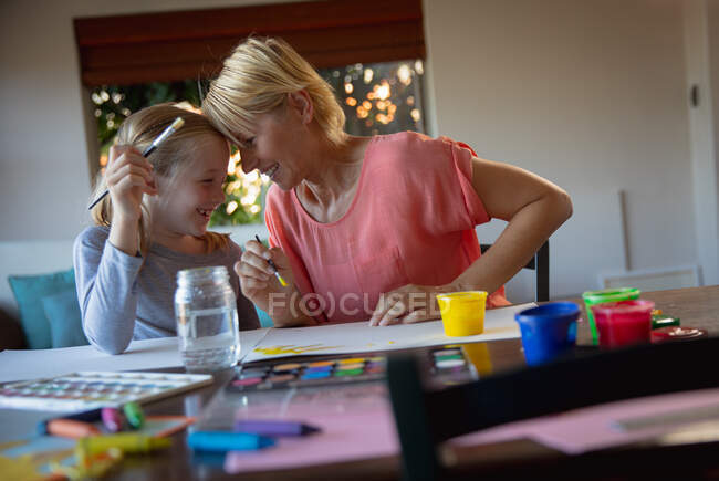 Vue de face d'une femme caucasienne profitant du temps en famille avec sa fille à la maison ensemble, assise à une table dans le salon, peignant, souriant et se regardant — Photo de stock